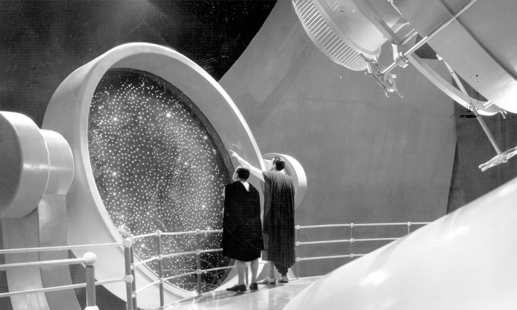 Two men in capes look at stars on a large screen