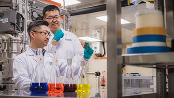 Two scientists in a lab looking at a black solar cell in front of four conical flasks full of colorful liquid