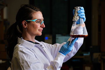 Woman in safety glasses holds up flask of fungus